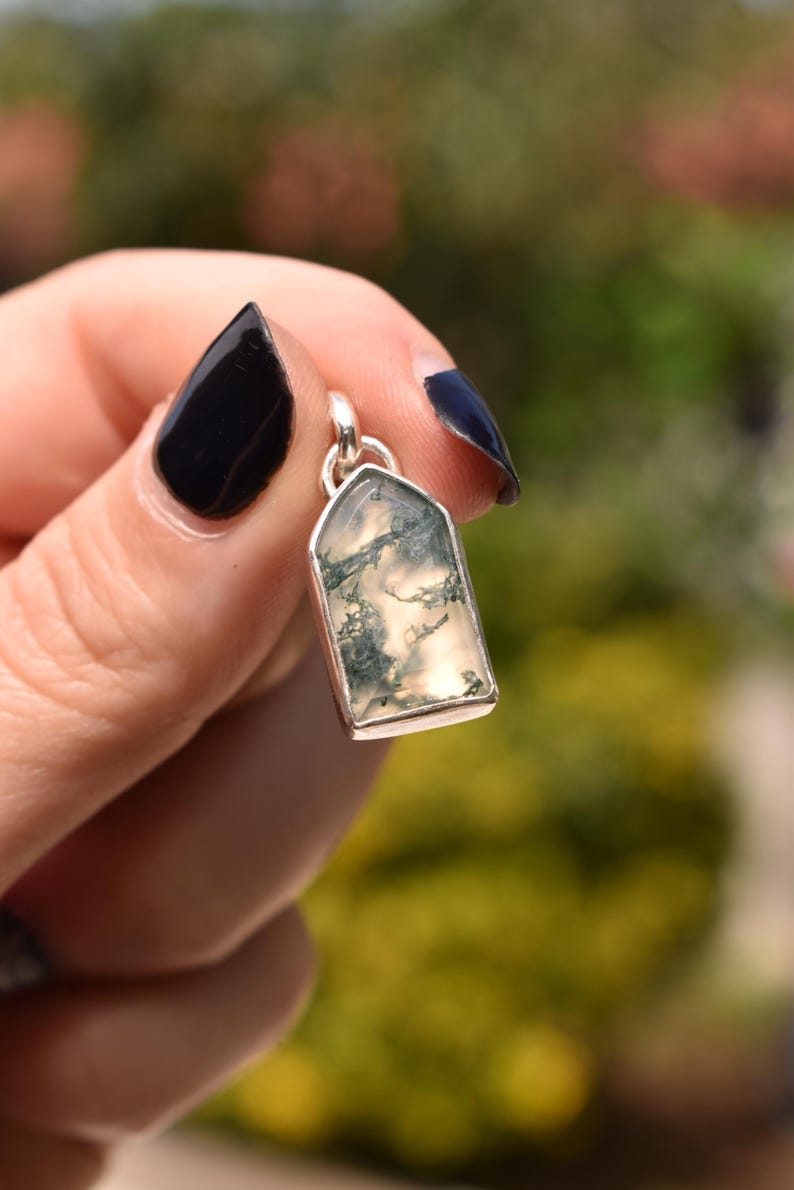 May include: A pendant featuring a translucent, shield-shaped moss agate stone with dark green, fern-like inclusions. The stone is encased in a polished silver-colored metal bezel setting with a small circular bail at the top for attachment.