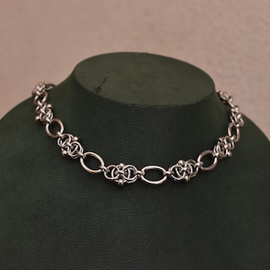 May include: A silver necklace with a detailed chain pattern. The necklace is composed of oval links and decorative, clustered components. It is presented on a dark green mannequin, set against a plain backdrop.