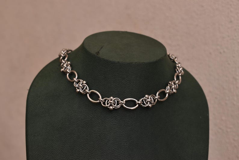May include: A silver necklace with a detailed chain pattern. The necklace is composed of oval links and decorative, clustered components. It is presented on a dark green mannequin, set against a plain backdrop.