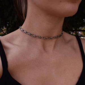 May include: A silver choker necklace with a chain-link design. The necklace has small, decorative elements evenly spaced along the chain. The choker is worn around the neck. The person is wearing a black top.