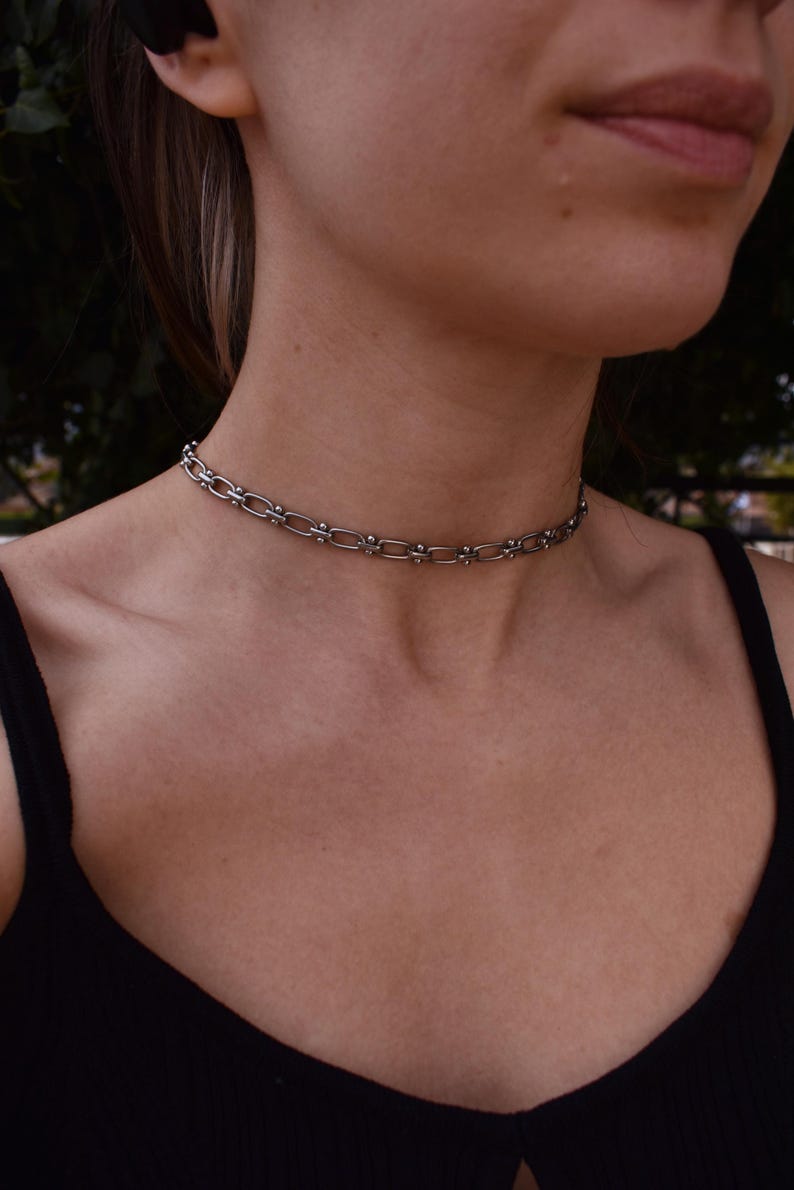 May include: A silver choker necklace with a chain-link design. The necklace has small, decorative elements evenly spaced along the chain. The choker is worn around the neck. The person is wearing a black top.
