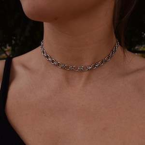 May include: A silver-coloured choker necklace with a chain-link design. The necklace is made of metal and sits close to the neck. The links are oval-shaped and connected by smaller, circular links. The background is blurred.