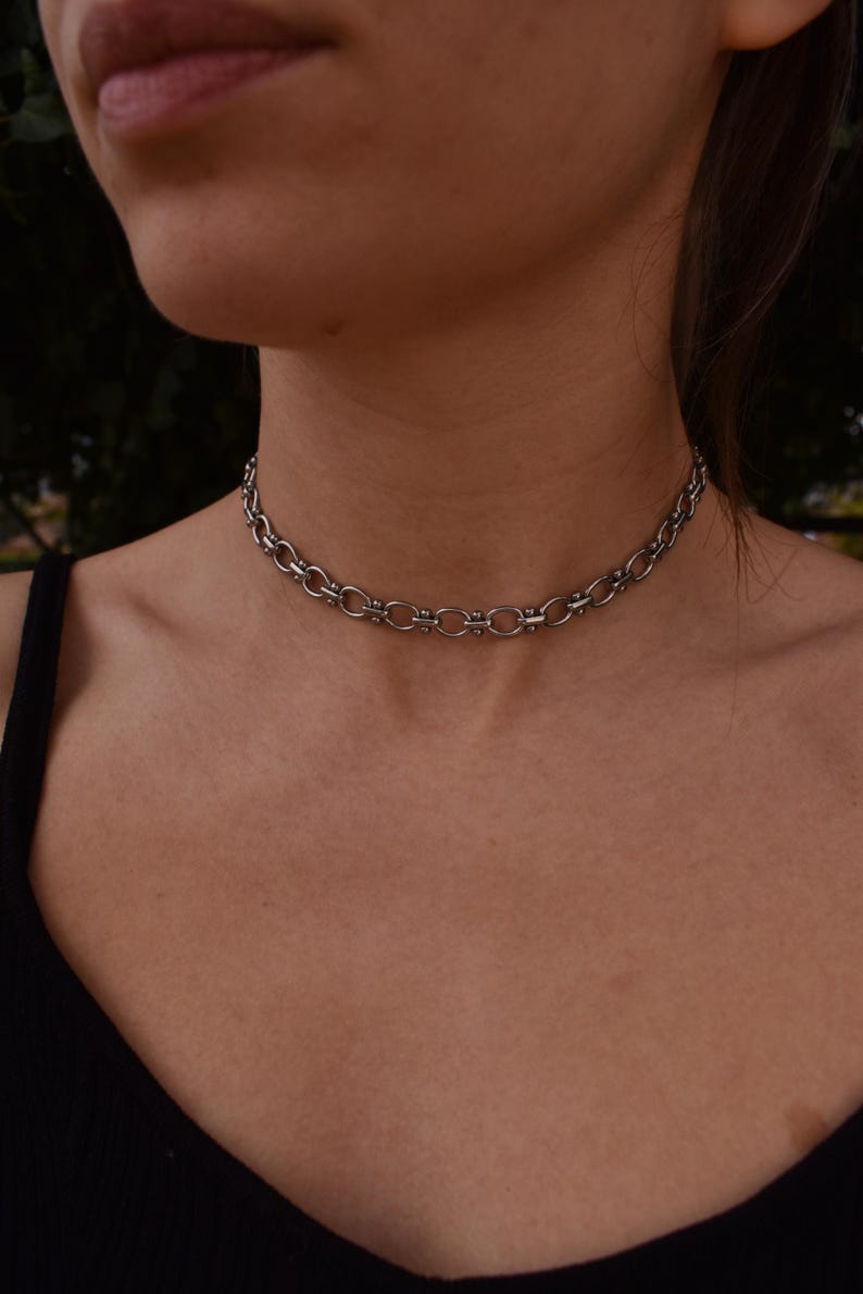 May include: A silver-coloured choker necklace with a chain-link design. The necklace is made of metal and sits close to the neck. The links are oval-shaped and connected by smaller, circular links. The background is blurred.