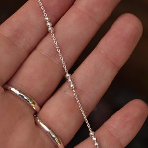 May include: A delicate silver chain necklace featuring small, evenly spaced metallic beads along its length. The chain is constructed from fine, polished silver links that catch the light, creating a subtle and refined appearance for this jewelry piece.