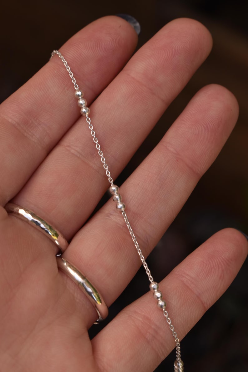 May include: A delicate silver chain necklace featuring small, evenly spaced metallic beads along its length. The chain is constructed from fine, polished silver links that catch the light, creating a subtle and refined appearance for this jewelry piece.