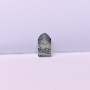 May include: A polished, tombstone-shaped moss agate gemstone featuring a translucent, milky white base with intricate, dark green, fern-like dendritic inclusions scattered throughout the stone, creating a unique, natural pattern within the flat surface.
