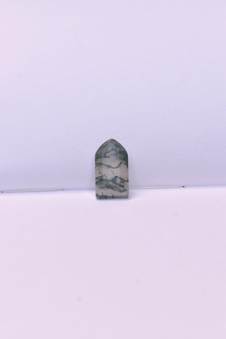 May include: A polished, tombstone-shaped moss agate gemstone featuring a translucent, milky white base with intricate, dark green, fern-like dendritic inclusions scattered throughout the stone, creating a unique, natural pattern within the flat surface.
