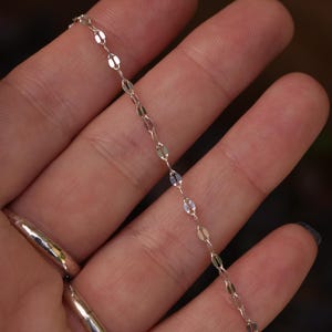 May include: A delicate silver chain necklace featuring small, flat, oval-shaped links that create a shimmering, reflective surface. The chain is composed of repeating metallic segments connected by tiny circular jump rings, forming a continuous, slender, and lightweight jewelry piece.
