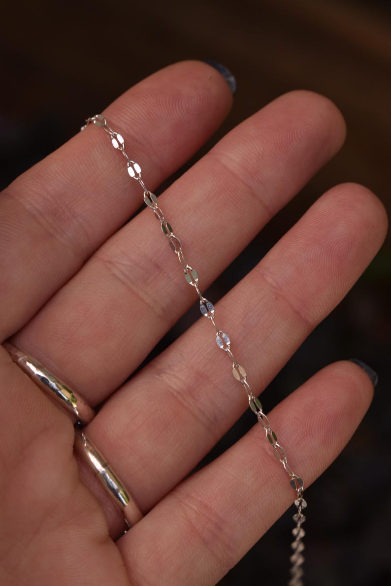 May include: A delicate silver chain necklace featuring small, flat, oval-shaped links that create a shimmering, reflective surface. The chain is composed of repeating metallic segments connected by tiny circular jump rings, forming a continuous, slender, and lightweight jewelry piece.