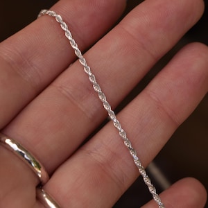 May include: A thin, silver-colored rope chain necklace featuring a twisted, spiraling link design. The metallic chain has a bright, reflective finish and a uniform, continuous texture throughout its length, showcasing a classic, simple jewelry style.