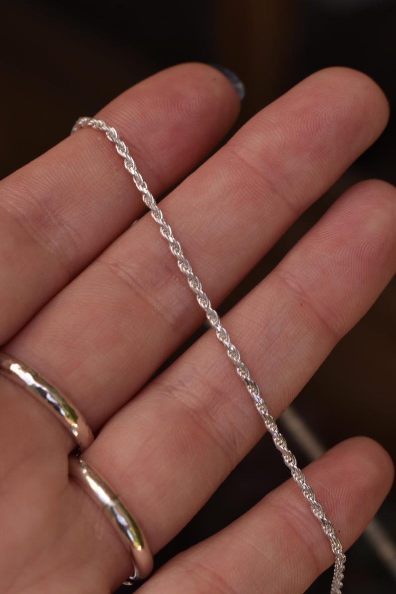 May include: A thin, silver-colored rope chain necklace featuring a twisted, spiraling link design. The metallic chain has a bright, reflective finish and a uniform, continuous texture throughout its length, showcasing a classic, simple jewelry style.