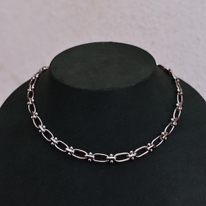 May include: A silver-toned necklace featuring an oval link chain design. The necklace is presented on a black mannequin bust against a neutral backdrop. The chain links are joined by small, round beads.