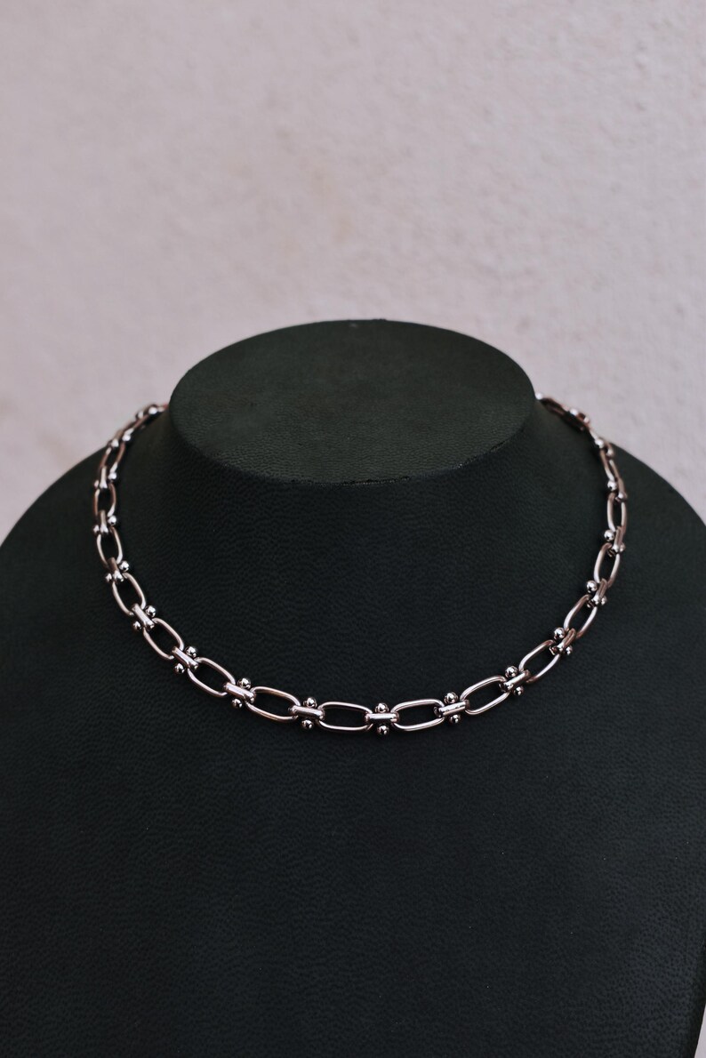 May include: A silver-toned necklace featuring an oval link chain design. The necklace is presented on a black mannequin bust against a neutral backdrop. The chain links are joined by small, round beads.