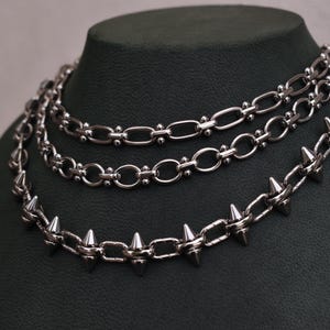 May include: Three silver necklaces on a dark green display. The top two have chain links, the bottom one has spikes. The necklaces are layered. The chains are made of metal and are displayed on a dark green mannequin.