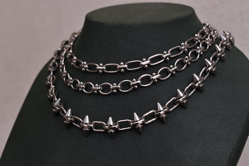 May include: Three silver necklaces on a dark green display. The top two have chain links, the bottom one has spikes. The necklaces are layered. The chains are made of metal and are displayed on a dark green mannequin.