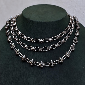 May include: Three silver necklaces on a dark green display. The necklaces feature different chain designs, including one with spikes. The chains are made of a shiny metal and are layered. The display is a dark green colour.
