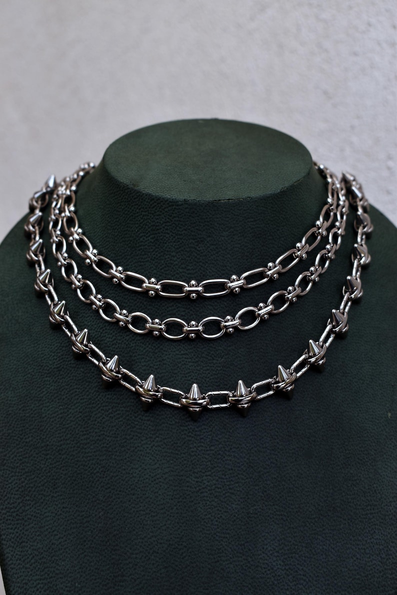 May include: Three silver necklaces on a dark green display. The necklaces feature different chain designs, including one with spikes. The chains are made of a shiny metal and are layered. The display is a dark green colour.