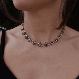 May include: A silver choker necklace with a chain design and evenly spaced, pointed spikes. The necklace is worn around the neck. The image is a close-up shot.