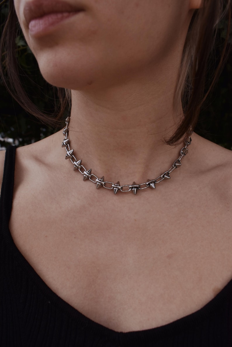 May include: A silver choker necklace with a chain design and evenly spaced, pointed spikes. The necklace is worn around the neck. The image is a close-up shot.