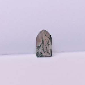 May include: A polished moss agate gemstone cut into a pointed arch shape. The stone features a translucent, pale pinkish-beige base color with dark green, branch-like dendritic inclusions spreading across the surface in a natural pattern.