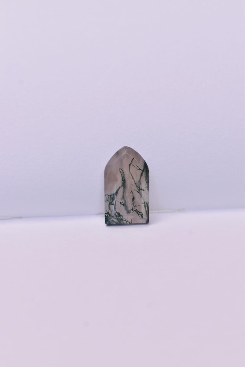 May include: A polished moss agate gemstone cut into a pointed arch shape. The stone features a translucent, pale pinkish-beige base color with dark green, branch-like dendritic inclusions spreading across the surface in a natural pattern.