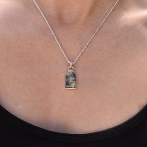 May include: A silver rope chain necklace featuring a rectangular pendant with an arched top. The pendant contains a translucent stone with dark, dendritic, tree-like inclusions that resemble a landscape scene set within a polished silver frame.