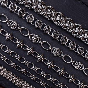 May include: A selection of silver metal chains in diverse designs, including spiked, oval link, and woven styles. The chains are presented on a dark, textured surface, highlighting their varied textures and designs. The chains are in various lengths.