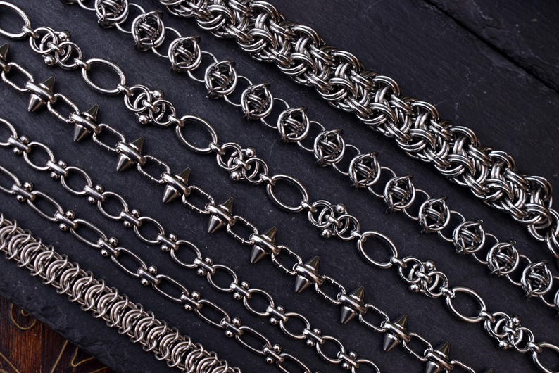 May include: A selection of silver metal chains in diverse designs, including spiked, oval link, and woven styles. The chains are presented on a dark, textured surface, highlighting their varied textures and designs. The chains are in various lengths.