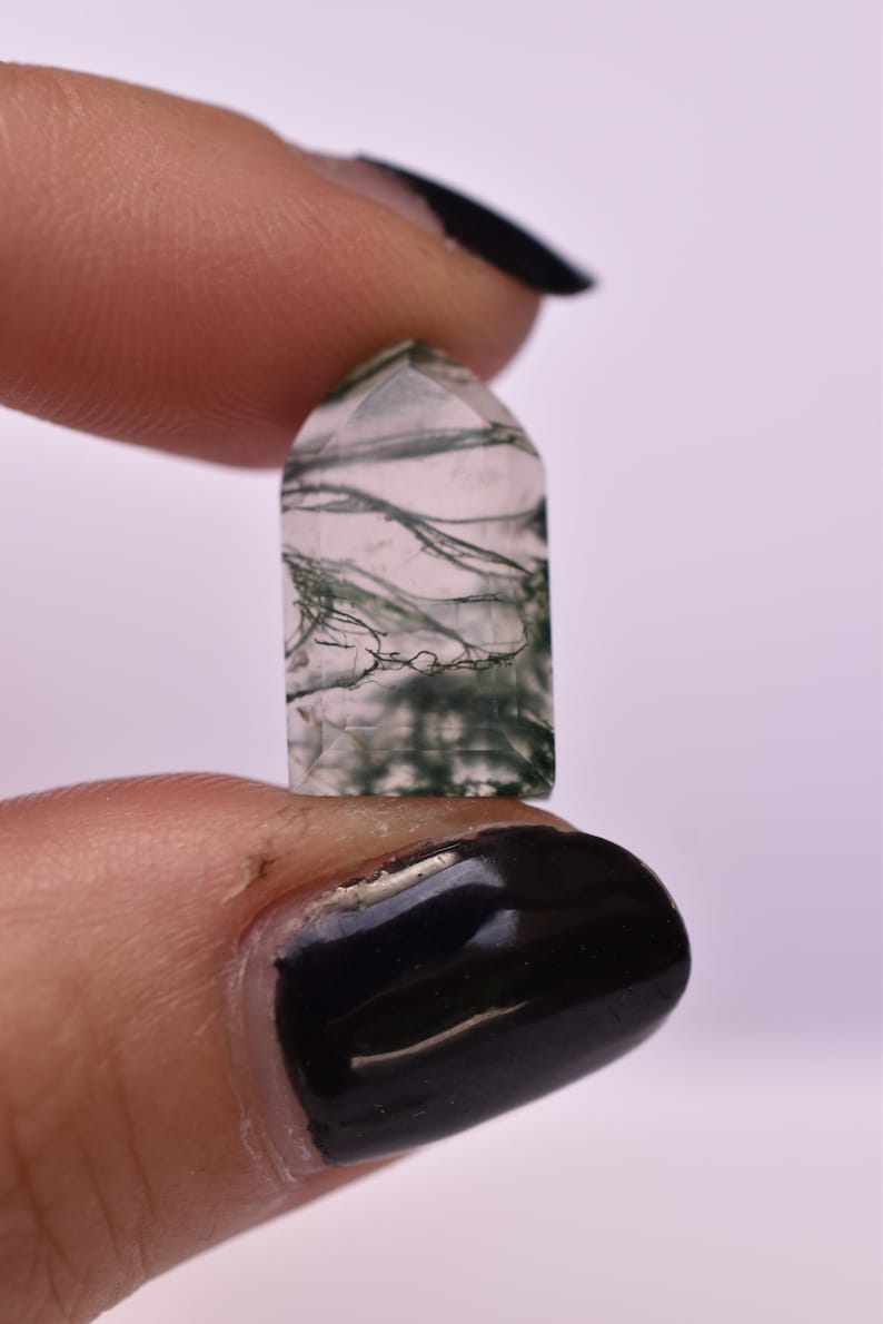 May include: A small, polished, semi-translucent moss agate gemstone shaped like a tombstone or arch. The stone features a clear, pale base with intricate, dark green, fern-like dendritic inclusions that spread throughout the interior of the crystal.