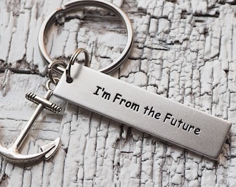 Op de afbeelding: Een zilverkleurige sleutelhanger met een rechthoekige metalen hanger waarop de tekst I'm From the Future is gestempeld, inclusief een kleine ankerbedel bevestigd aan een ronde splitring van glad, geborsteld metaal.