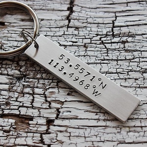 May include: A silver keychain with a rectangular tag engraved with the coordinates "53.5571°N 113.4968°W".