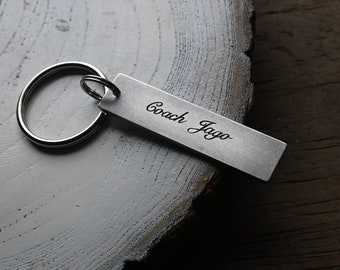 Op de afbeelding: Zilveren sleutelhanger met een rechthoekig label met de tekst "Coach Jago" gegraveerd.