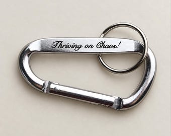 Op de afbeelding: Een zilverkleurige metalen karabijnhaak-sleutelhanger met een veerbelaste sluiting en een ronde splitring. De platte zijde van de karabijnhaak is gegraveerd met de zwarte tekst "Thriving on Chaos!" in een cursief lettertype, een functioneel accessoire.