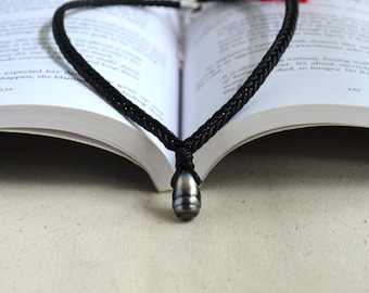 May include: A black braided cord necklace featuring a single teardrop-shaped dark gray pearl pendant. The cord is knotted securely around the top of the pearl, creating a simple and clean design that rests against the open pages of a book.