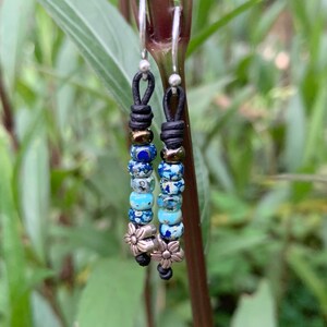 May include: A pair of dangle earrings with silver hoops and black leather cords. The cords are strung with blue, turquoise, and gold-colored beads and silver flower charms.