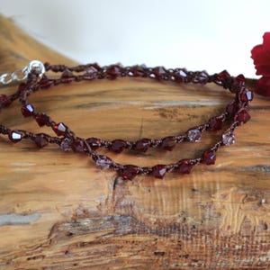 Könnte beinhalten: Eine handgefertigte Halskette mit einem doppelreihigen Design. Die Halskette hat dunkelrote Perlen und einen silbernen Verschluss. Der Schmuck wird auf einer Holzoberfläche mit roten Blumen im Hintergrund präsentiert.