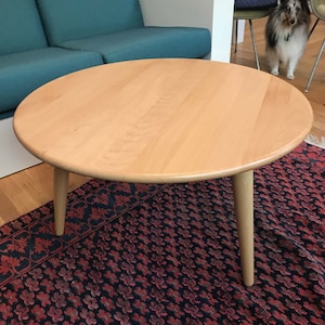 May include: A round wooden coffee table with a light wood finish and three legs. The table is sitting on a red and black patterned rug.