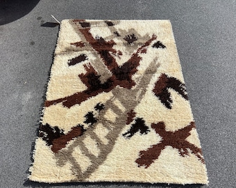 May include: A beige rug with a geometric pattern in brown and black. The rug has a shaggy texture and is made of wool.