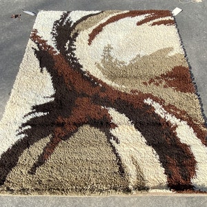 May include: A vintage shag rug with a brown, beige, and white abstract design. The rug has a long, shaggy pile and is in good condition.