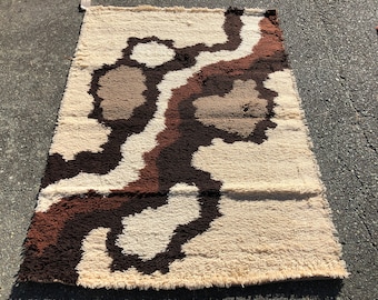 May include: A cream-coloured rug with a brown and beige abstract design. The rug has a shaggy texture and a fringe border.