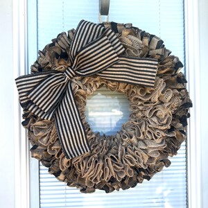 May include: A round wreath with a burlap base and a large black and beige striped bow. The wreath is made of ruffled burlap and has a neutral color palette, suitable for various home decor styles.