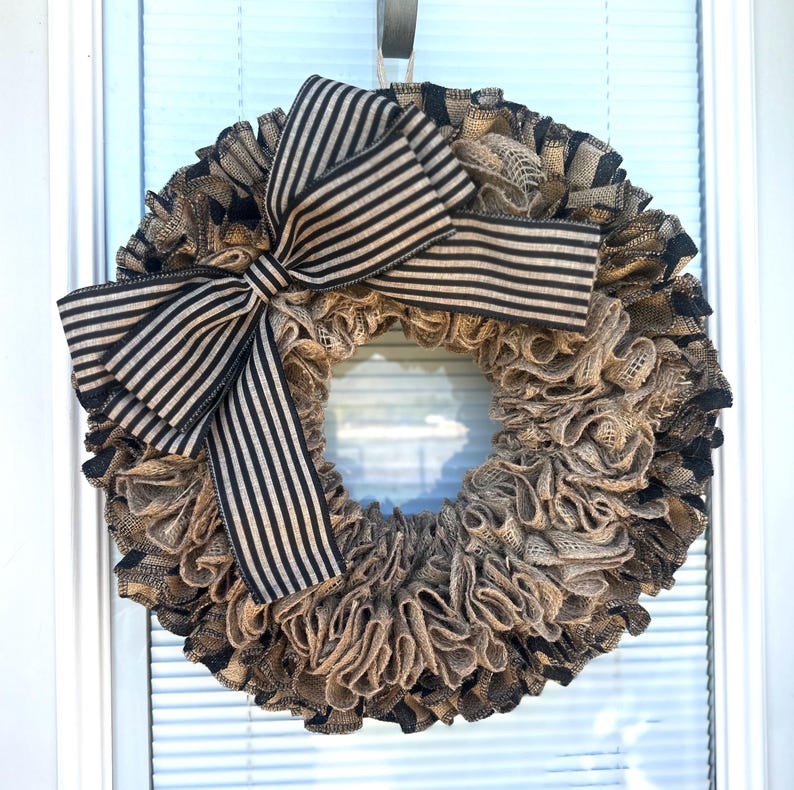 May include: A round wreath with a burlap base and a large black and beige striped bow. The wreath is made of ruffled burlap and has a neutral color palette, suitable for various home decor styles.
