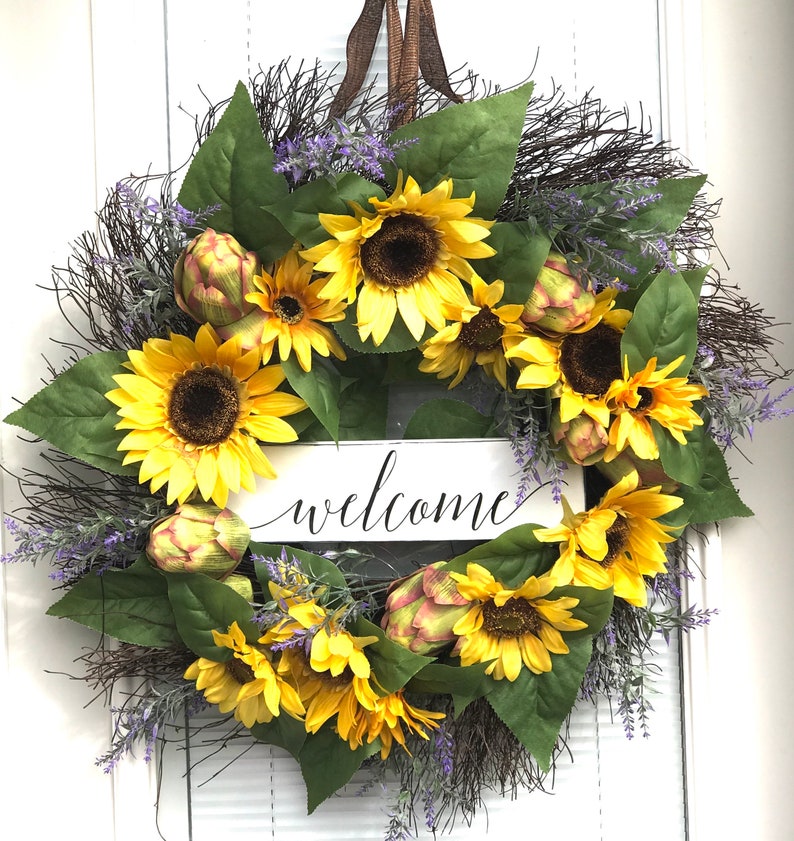 Pu&ograve; includere: Una ghirlanda di girasoli con un cartello di benvenuto. La ghirlanda &egrave; fatta di girasoli artificiali, verde e fiori viola. Il cartello &egrave; bianco con lettere nere.