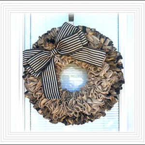 May include: A decorative wreath with a ruffled burlap design in shades of tan and black. A large bow with black and white stripes is centered at the top. The wreath is hanging on a white window.