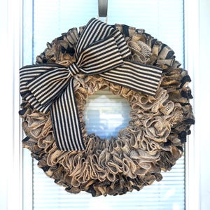 May include: A round wreath made of layered burlap in shades of tan and black. A large bow with black and tan stripes is centered at the top. The wreath is hanging on a white door.