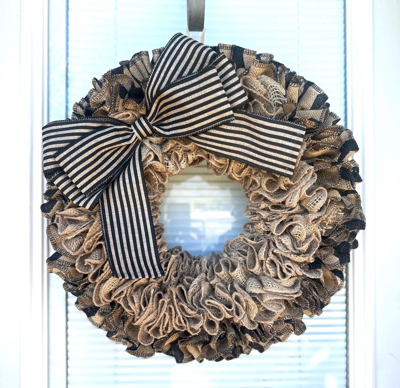 May include: A round wreath made of layered burlap in shades of tan and black. A large bow with black and tan stripes is centered at the top. The wreath is hanging on a white door.