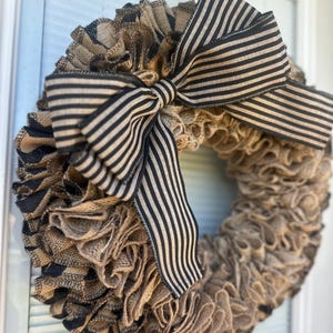 May include: A handmade burlap wreath with a large black and white striped bow. The wreath is made of layered burlap in shades of brown and tan, with accents of black burlap. This decorative wreath is perfect for a front door or wall.