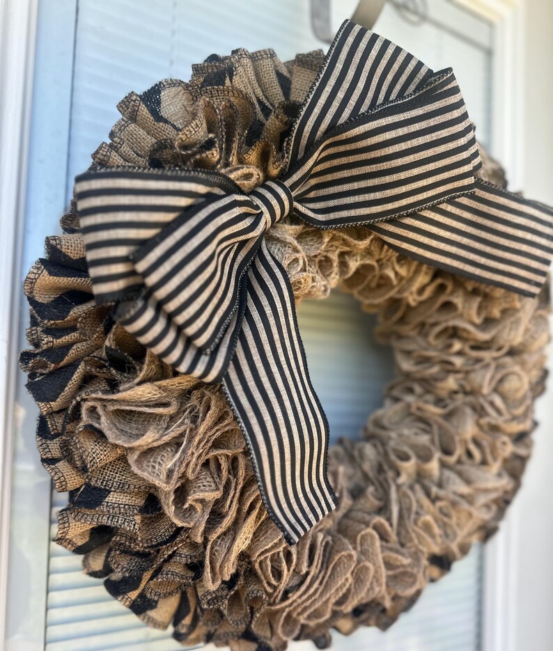 May include: A handmade burlap wreath with a large black and white striped bow. The wreath is made of layered burlap in shades of brown and tan, with accents of black burlap. This decorative wreath is perfect for a front door or wall.
