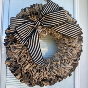 May include: A rustic burlap wreath with a large bow. The wreath is made of layered, ruffled burlap in shades of brown and tan. A black and cream striped bow is centered at the top, adding a contrasting accent. This decorative wreath is perfect for a front door.