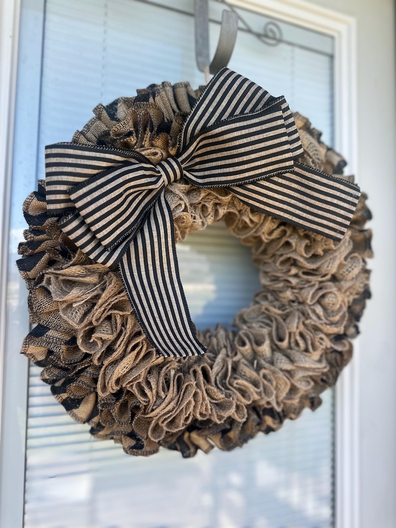 May include: A rustic burlap wreath with a large bow. The wreath is made of layered, ruffled burlap in shades of brown and tan. A black and cream striped bow is centered at the top, adding a contrasting accent. This decorative wreath is perfect for a front door.
